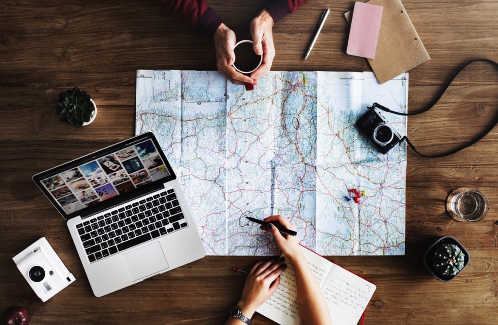 Dos personas viendo un mapa, hay una laptop, libreta, cámara y plantas